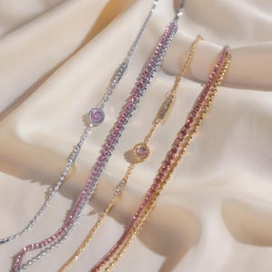 Op de afbeelding: Vier delicate armbanden in zilver- en goudtinten, versierd met roze en heldere edelstenen. Elke armband heeft een ander ontwerp, waaronder een rij kleine kralen en een rij grotere stenen. De armbanden worden gepresenteerd op een crèmekleurige stof.