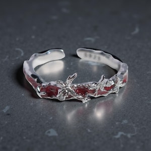 May include: A silver-colored adjustable ring with a red center and floral design. The ring has a textured, organic shape with small, clear stones and the text "S925" on the inside.