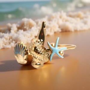 May include: A gold-colored ring featuring a seashell, a textured coral-like element, and a light blue starfish. The ring is set against a blurred beach background, with the ocean in the distance. The ring's design evokes a coastal theme.