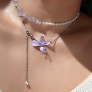 May include: A layered necklace featuring a delicate lavender flower pendant. The pendant is a four-petal design with silver-tone metal. The necklace also includes a strand of small pearls and a strand of light purple gemstones. A single pearl dangles from the pendant.