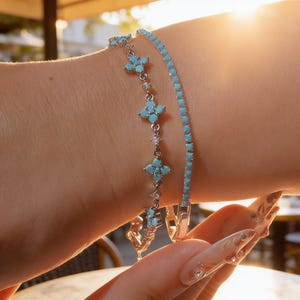 May include: Two silver-toned bracelets worn together. One bracelet features a repeating pattern of four-petaled flower motifs set with light blue stones and small clear crystals. The second bracelet is a thin, flexible chain of light blue stones.