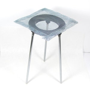 May include: A silver metal tripod stand with a wire mesh platform. The platform is square and has a round hole in the center.