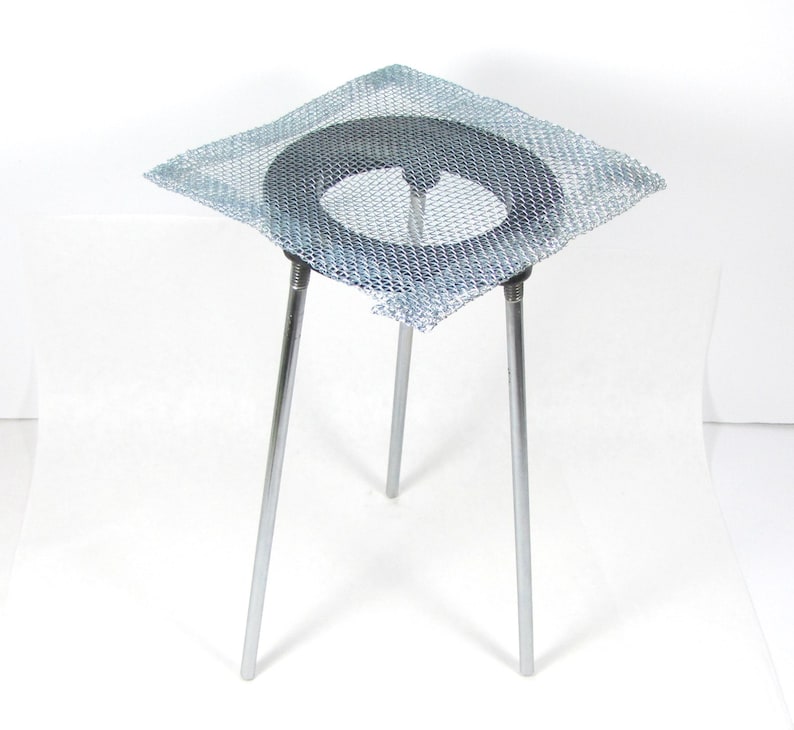 May include: A silver metal tripod stand with a wire mesh platform. The platform is square and has a round hole in the center.