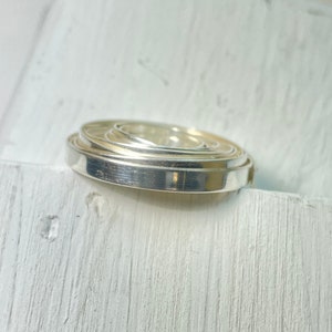 May include: A stack of silver-colored metal rings, each decreasing in size, creating a layered effect. The rings have a smooth, reflective surface, and are positioned on a white, textured surface. The overall aesthetic is minimalist and modern.