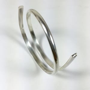 Może przedstawiać: Srebrna bransoletka o prostym, eleganckim wzorze. Bransoletka wykonana jest z jednego, ciągłego kawałka metalu skręconego w kształt spirali.
