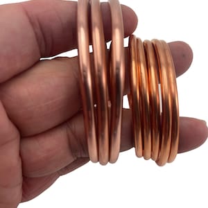 May include: A set of four copper bangles, two larger and two smaller, stacked on top of each other. The bangles are smooth and shiny, with a warm copper color.