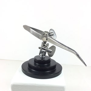 May include: A silver-toned, adjustable, third-hand soldering tool with a black base. The tool has a long, curved arm with a pair of tweezers at the end.