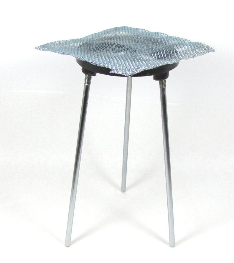 May include: A silver wire mesh sits on top of a black metal ring, which is supported by three silver metal legs. The legs are attached to the ring with screws. The wire mesh is used to support beakers or other lab equipment over a heat source.