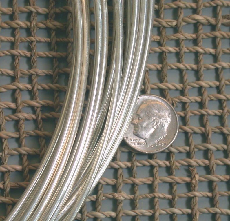 May include: A close-up of a bundle of silver wire, with a dime for scale. The wire is shiny and smooth, and it is arranged in a neat pile. The dime is positioned in the lower right corner of the image.