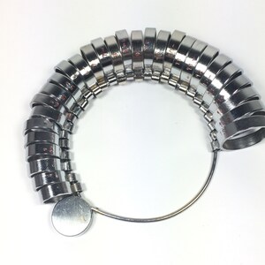May include: A silver ring sizer with a series of graduated rings, numbered 1 to 13, attached to a metal loop. The rings are arranged in a semi-circle shape.