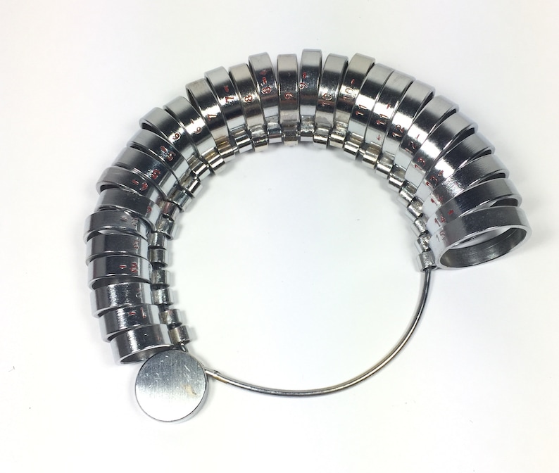May include: A silver ring sizer with a series of graduated rings, numbered 1 to 13, attached to a metal loop. The rings are arranged in a semi-circle shape.