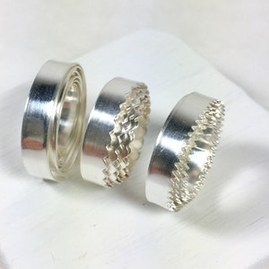 May include: Three silver rings with different designs. The ring on the left is a plain band, the middle ring has a wavy edge, and the ring on the right has a serrated edge.
