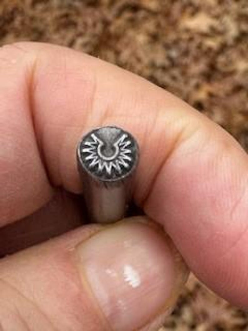 May include: Close-up of a metal stamp with a circular design featuring a stylized crescent moon and radiating lines.