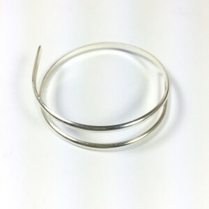 Może przedstawiać: Srebrna bransoletka typu bangle o prostym, minimalistycznym designie. Bransoletka wykonana jest z dw&oacute;ch cienkich, okrągłych drut&oacute;w, kt&oacute;re są ze sobą splecione.