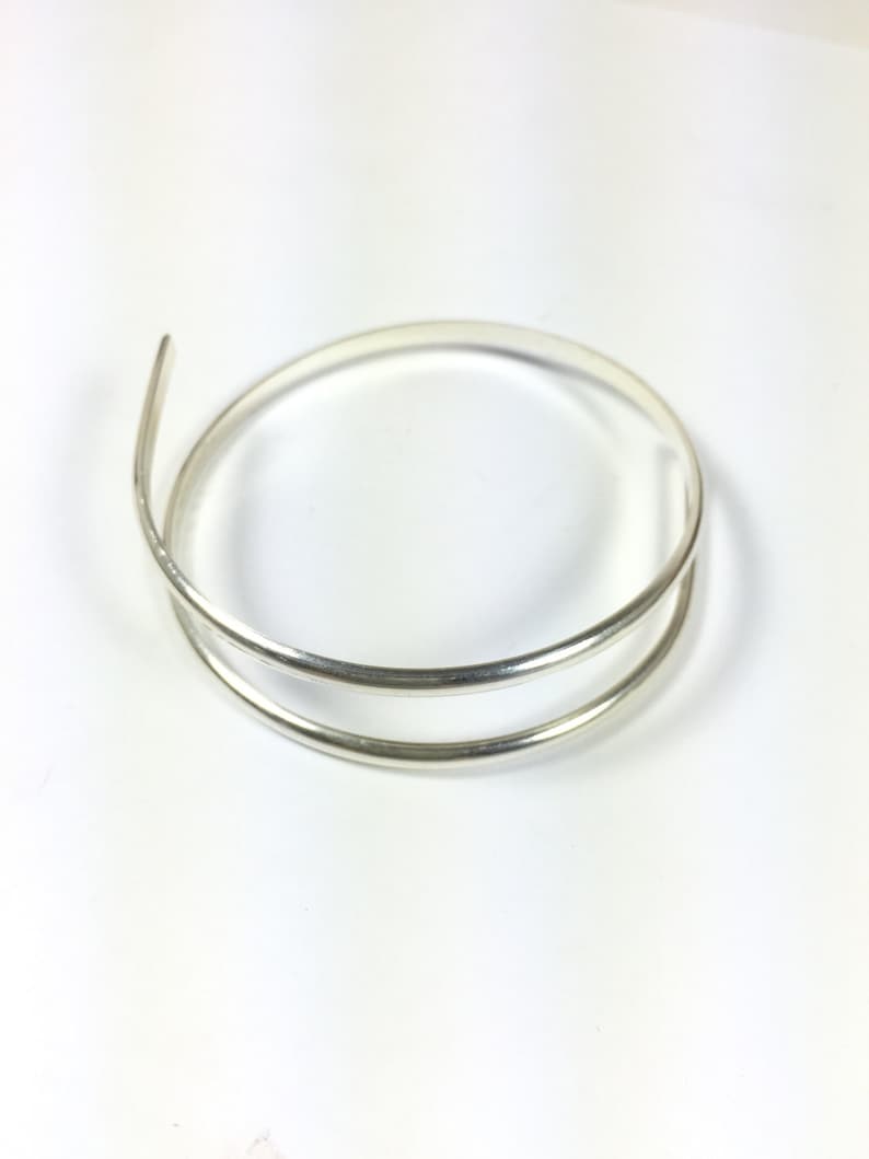 Może przedstawiać: Srebrna bransoletka typu bangle o prostym, minimalistycznym designie. Bransoletka wykonana jest z dw&oacute;ch cienkich, okrągłych drut&oacute;w, kt&oacute;re są ze sobą splecione.