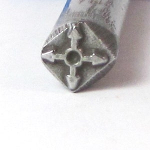 K&ouml;nnte beinhalten: Nahaufnahme eines silbernen Metallstempels mit einem rautenf&ouml;rmigen Design. Das Design zeigt vier Pfeile, die in verschiedene Richtungen zeigen, mit einem kleinen Kreis in der Mitte.
