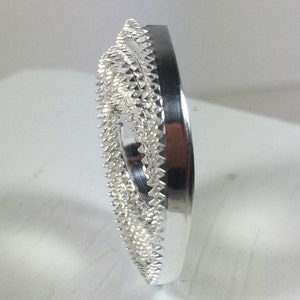 May include: A silver-colored metal ring with a decorative, textured design. The ring has a circular shape and a smooth, polished finish.