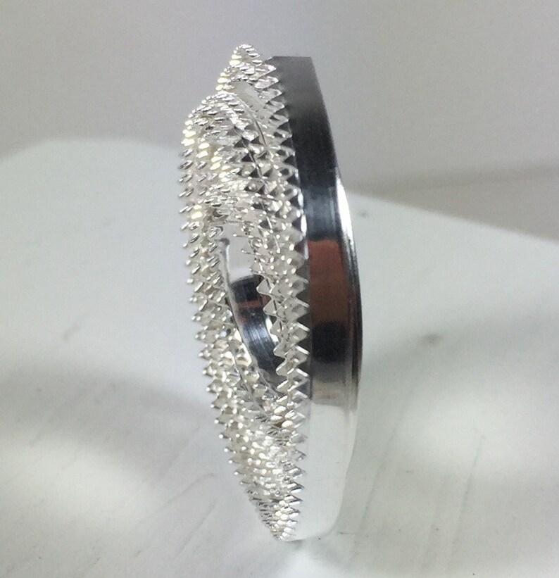 May include: A silver-colored metal ring with a decorative, textured design. The ring has a circular shape and a smooth, polished finish.
