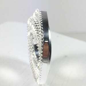 May include: A silver ring with a detailed, intricate design. The ring is a wide band with a textured surface.