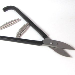 May include: Black handled shears with silver blades. The shears are open and there are two silver metal coils on the table.