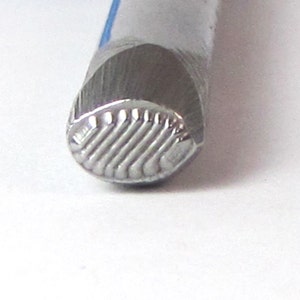 May include: Close-up of a silver metal stamp with a textured, ridged design. The stamp is used for creating impressions in leather or metal.