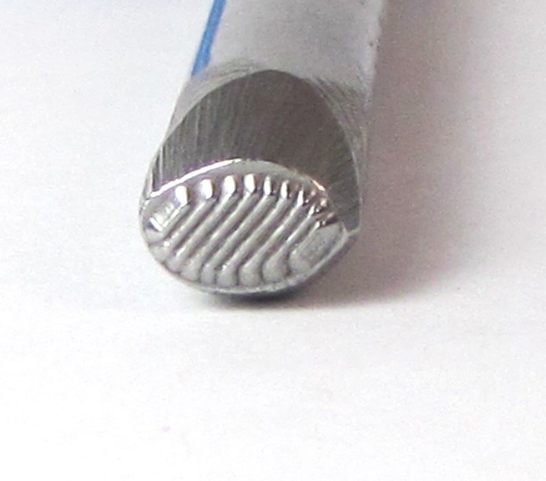 May include: Close-up of a silver metal stamp with a textured, ridged design. The stamp is used for creating impressions in leather or metal.