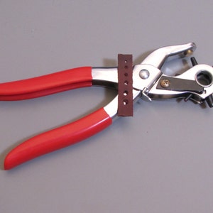 May include: A red handled leather punch tool with a silver metal frame and a brown leather guide. The tool has a circular punch with multiple holes.