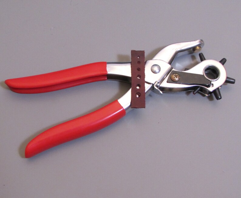 May include: A red handled leather punch tool with a silver metal frame and a brown leather guide. The tool has a circular punch with multiple holes.