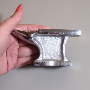 May include: A small, silver-colored metal anvil with a rounded top and a flat base. The anvil is about 4 inches tall and 3 inches wide.