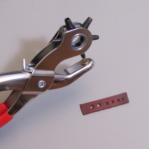 May include: A red-handled metal leather punch tool with a silver finish. The tool is used to create holes in leather. A brown leather strip with multiple holes is visible in the background.