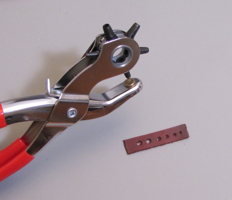 May include: A red-handled metal leather punch tool with a silver finish. The tool is used to create holes in leather. A brown leather strip with multiple holes is visible in the background.