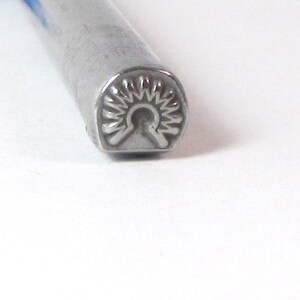 May include: A close-up of a silver metal stamping tool with a sunburst design. The tool is used for creating impressions in metal.