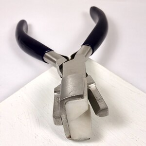May include: Jewelry-making pliers with a silver metal body and dark blue handles. The pliers have a white, rectangular tip. The tool is designed for bending and shaping metal wire and other jewelry components.