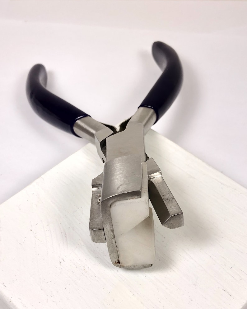 May include: Jewelry-making pliers with a silver metal body and dark blue handles. The pliers have a white, rectangular tip. The tool is designed for bending and shaping metal wire and other jewelry components.