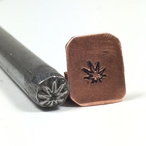 May include: A close-up of a metal stamp with a flower design. The stamp is used to create impressions in metal. The stamp is shown next to a piece of copper that has been stamped with the flower design.