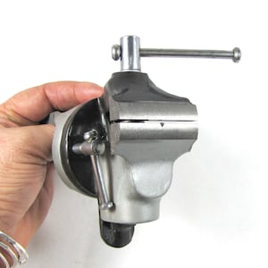 May include: A silver and black metal bench vise with a swivel base and a handle for tightening the jaws.