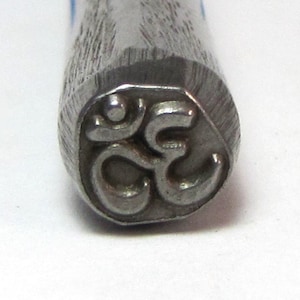 May include: Close-up of a metal stamp with the Om symbol engraved on the face. The stamp is silver in color and has a textured surface.