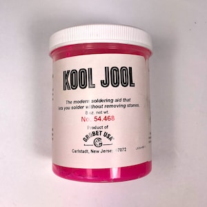 May include: A cylindrical container of Kool Jool soldering aid. The container is pink with a white lid and a label with black text that reads "KOOL JOOL". The label also states the product is 8 oz. net wt. and lists the manufacturer as Grobet USA.