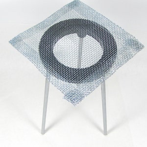 May include: A square wire mesh with a black ring in the center, supported by three metal legs. The mesh is designed to be used as a support for beakers or other laboratory equipment.