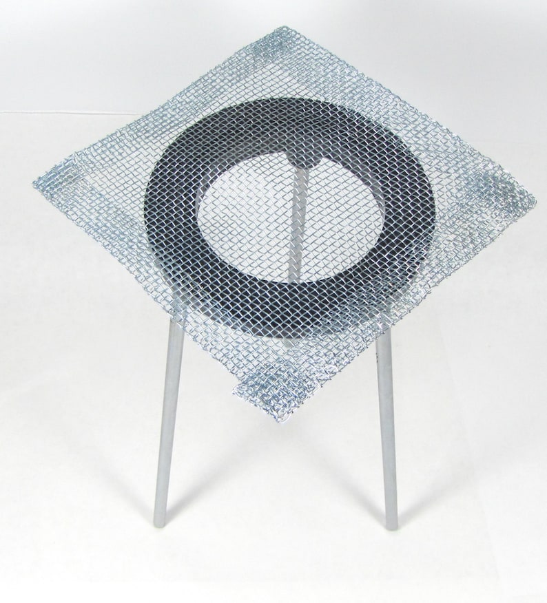 May include: A square wire mesh with a black ring in the center, supported by three metal legs. The mesh is designed to be used as a support for beakers or other laboratory equipment.