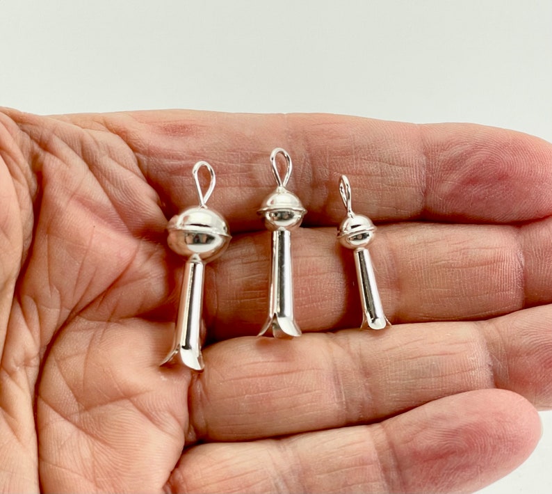 May include: Three silver pendant charms in the shape of a stylized flower with a round top. The charms are different sizes and are shown in a hand.