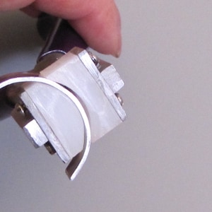 May include: A close-up of a silver metal ring bender tool with a white plastic insert. The tool is used to bend metal rings into different shapes.