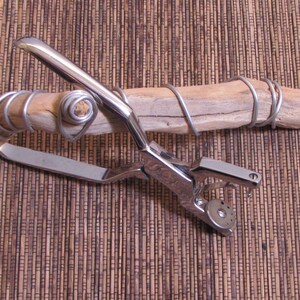 May include: A silver metal tool with a handle and a circular punch. The tool is resting on a piece of driftwood wrapped with silver wire.