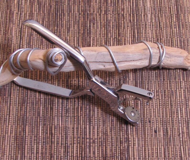 May include: A silver metal tool with a handle and a circular punch. The tool is resting on a piece of driftwood wrapped with silver wire.
