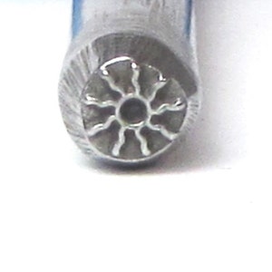 May include: Close-up of a metal stamp with a sun design. The stamp is silver and has a circular shape with eight rays emanating from the center.