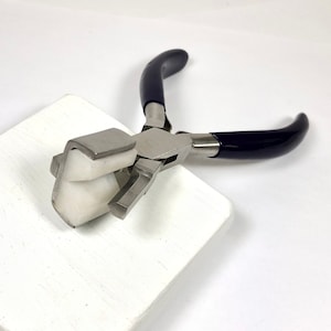 May include: A pair of jewelry pliers with a stainless steel body and dark blue handles. The pliers are resting on a white, textured surface. The pliers have a unique design, with a flat, white, protective jaw.