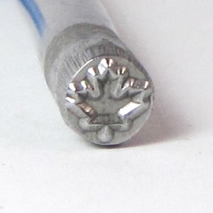 May include: Close-up of a silver metal stamp with a maple leaf design.