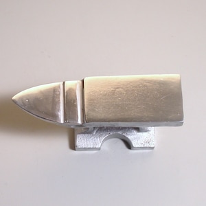 May include: A small, silver-colored metal anvil with a flat top and a rounded horn. The anvil is mounted on a base with a cutout in the front.