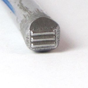 May include: Close-up of a metal stamping tool with a square head and three horizontal lines.
