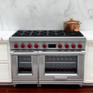 May include: A miniature silver-colored kitchen range with a black stovetop grate, ten red control knobs, and two glass-front oven doors. A small copper-colored pot with a lid sits on the right side of the stovetop surface of the appliance.